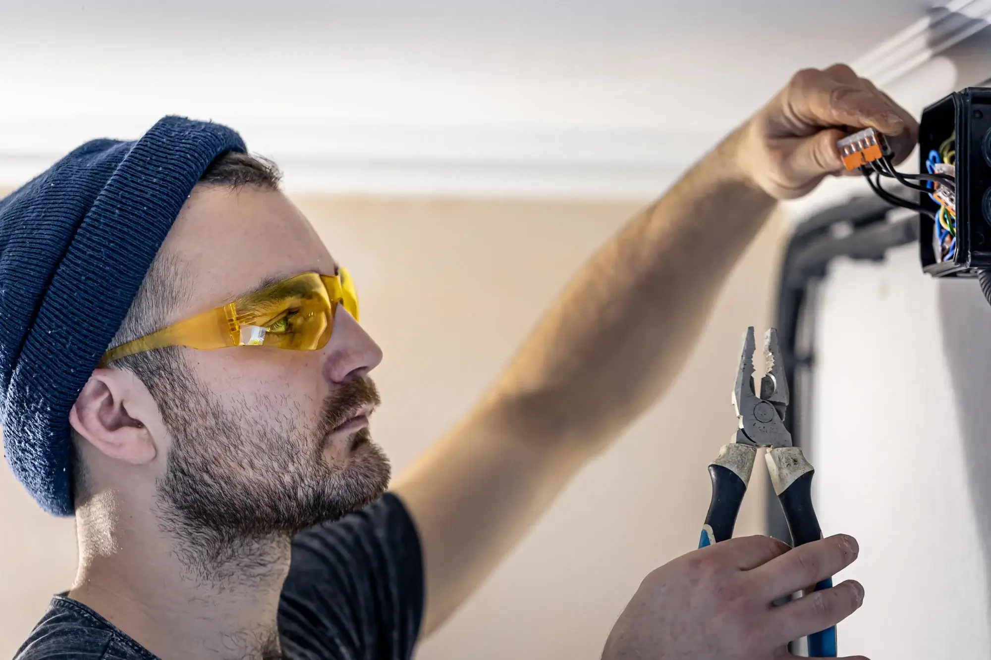 Electrician in beanie and yellow safety glasses using pliers on a wall-mounted junction box fitted with WAGO push-in connectors