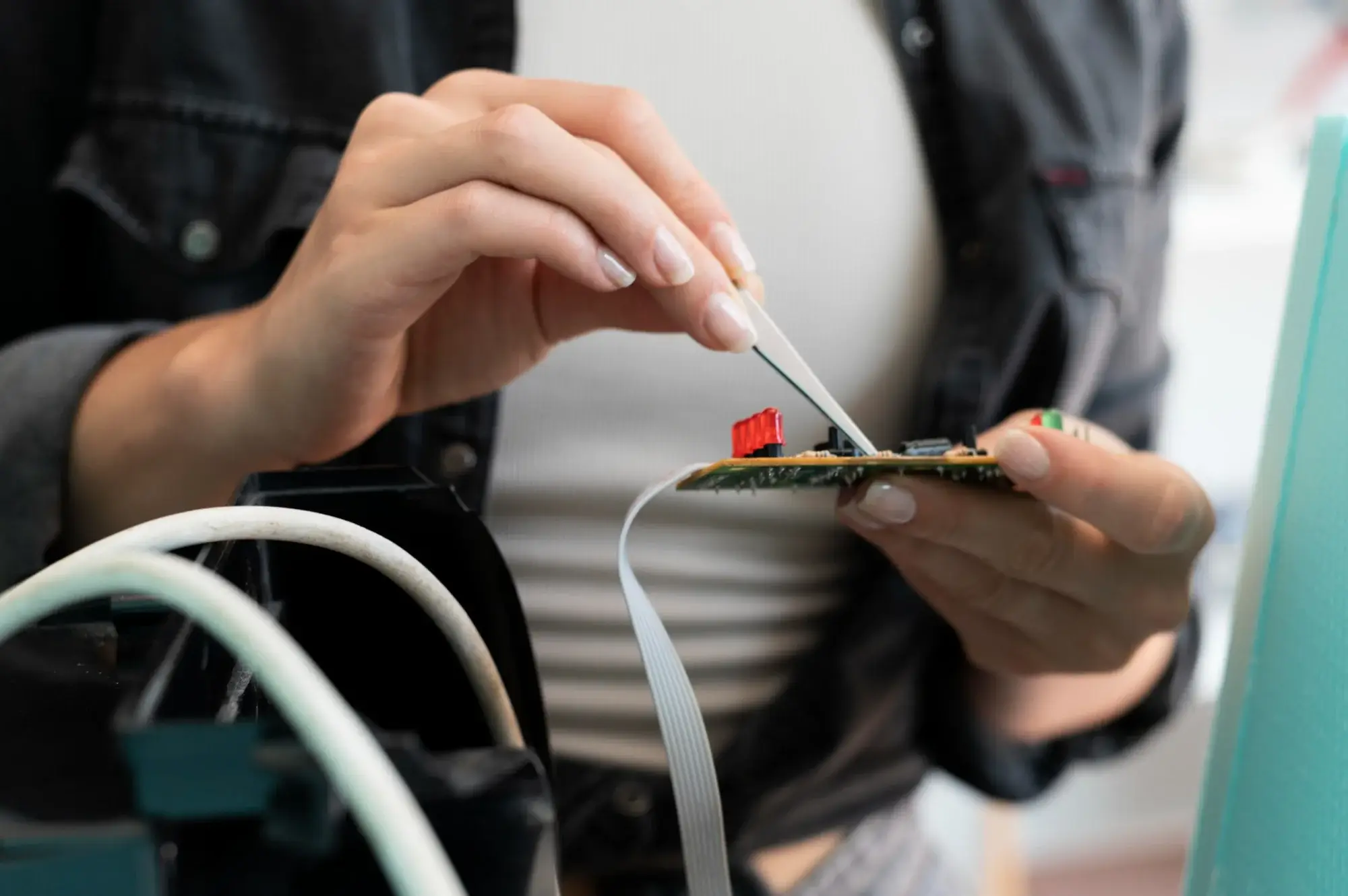 Close-up of a technician using fine tweezers to work on a small printed circuit board with attached ribbon cable