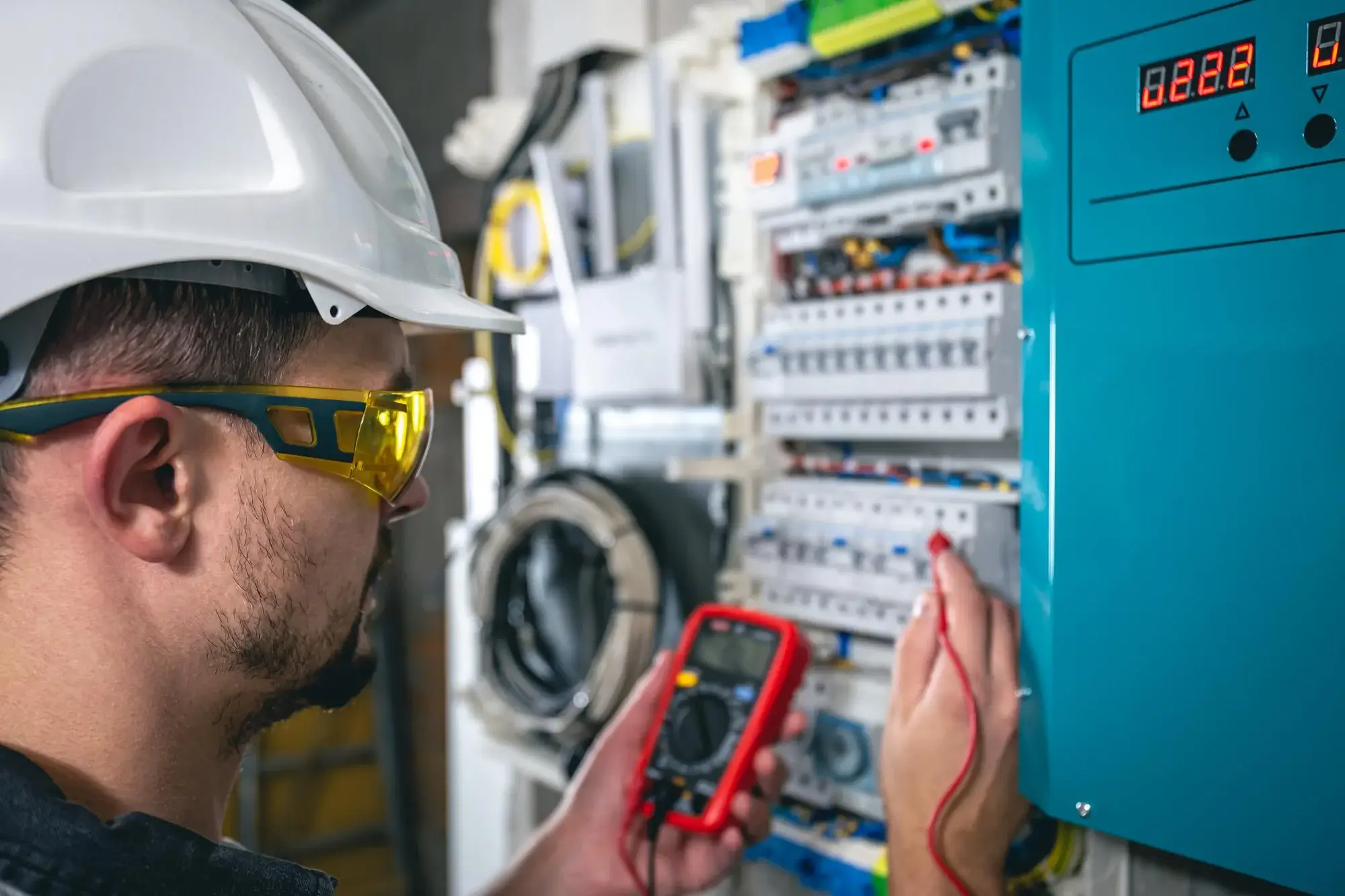 Electrician in white hard hat and yellow safety glasses using a red multimeter to test a control panel beside a washing machine