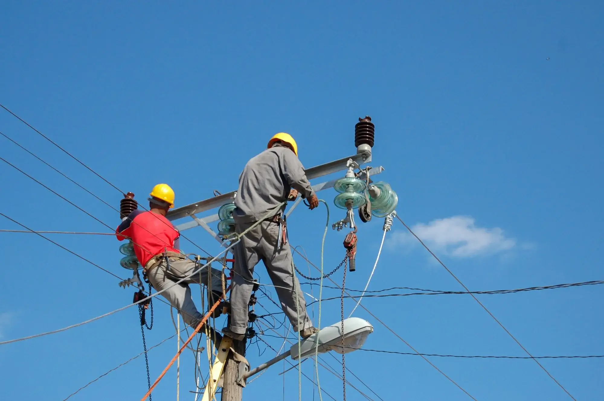 Two overhead linemen in hard hats and harnesses working at the top of a wooden power pole with insulators against a blue sky