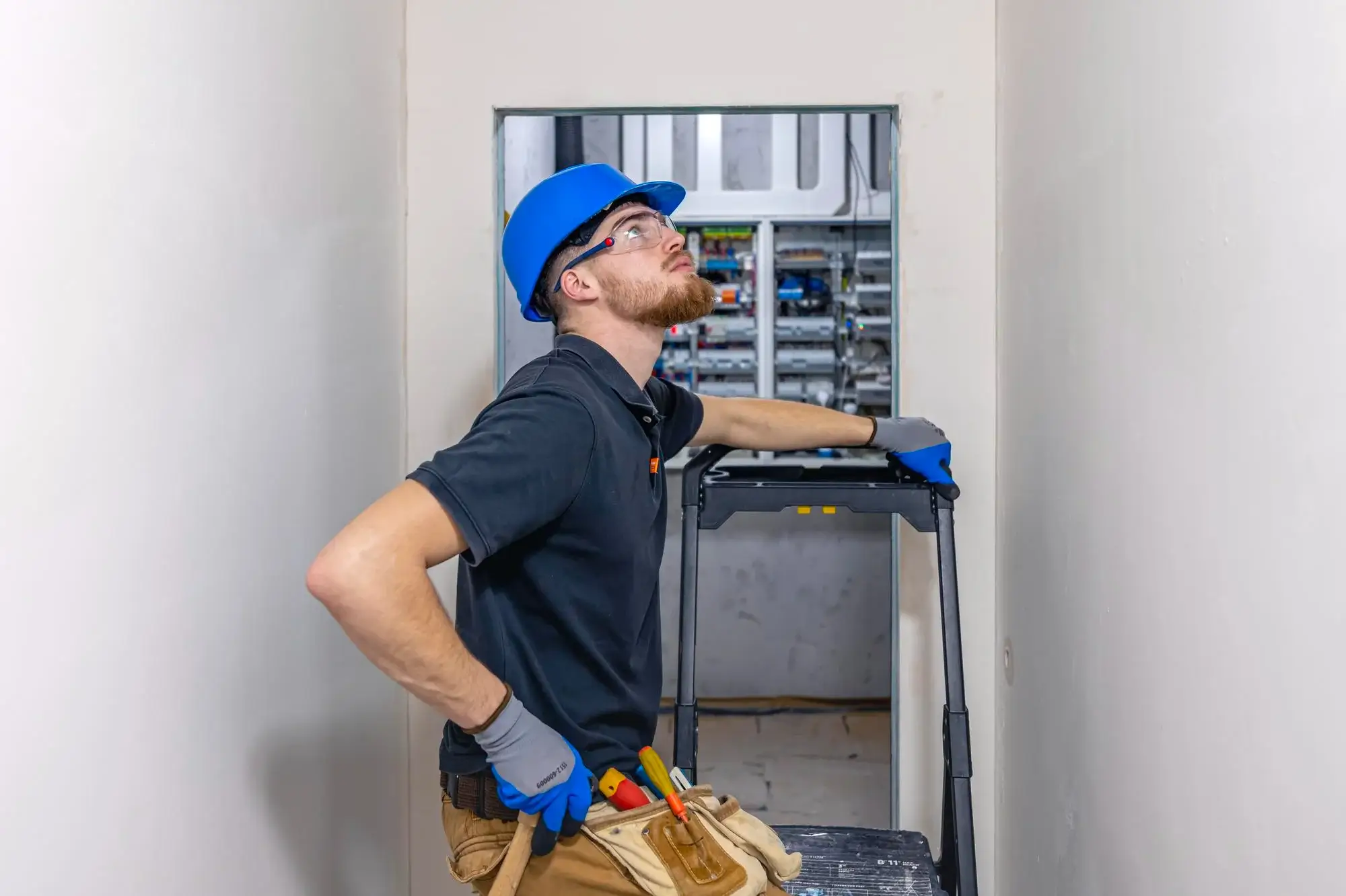 Electrician in blue hard hat standing with a stepladder while looking up at a ceiling fixture beside an opened distribution panel