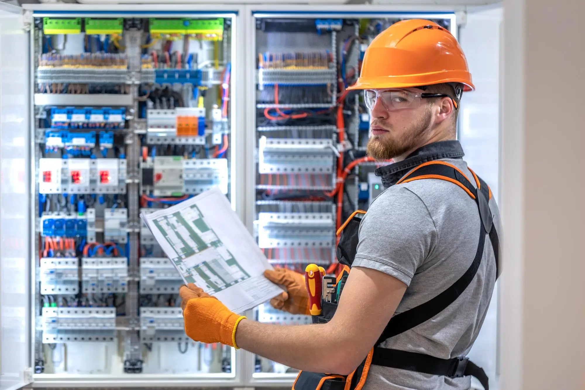 Electrician in orange hard hat and safety glasses reviewing a wiring schematic at an open industrial switchboard