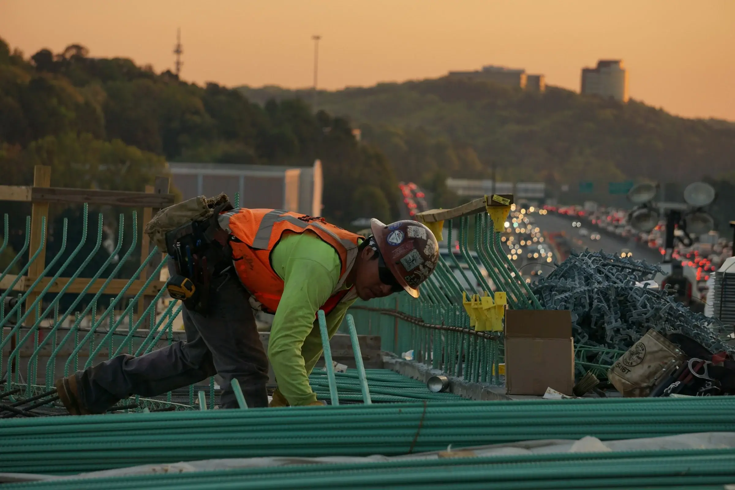 Construction worker in orange high-vis vest and hard hat tying rebar at sunset on a highway bridge deck
